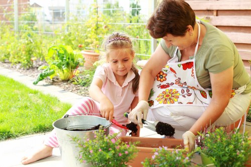 A woman and a young girl are engaged in gardening activities in a backyard garden, with the woman wearing an apron and gardening gloves, and the girl sitting on the grass wearing a pink striped outfit. They are planting or tending to potted plants in a rectangular wooden planter box, which is positioned on a paved outdoor area adjacent to a lush green lawn. The garden background features various plants, shrubs, and a wire fence, with trees and other garden elements visible in the distance. The scene is illuminated by natural daylight, highlighting the vibrant green foliage and colorful flowering plants. The setting appears well-maintained and reflects typical outdoor gardening work common in residential gardens, aligned with gardening and lawn care services offered by Lawn Mowing Lambeth in the Lambeth area, postcode well associated with London boroughs, creating a natural connection to local outdoor maintenance services.