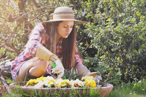 In a well-maintained garden, a young woman wearing a large straw sunhat, a red plaid shirt, and gardening gloves is tending to a flower bed. She is carefully planting or adjusting yellow, white, and purple flowers in a circular flower bed bordered by wooden edging. Surrounding her, the garden features a lush, green lawn with dense shrubbery and mature trees in the background, providing a natural and vibrant outdoor environment. The sunlight filters through the foliage, indicating a bright day with clear weather. This scene reflects attentive garden care typical of professional landscaping services in Lambeth, highlighting the importance of detailed plant maintenance and outdoor aesthetic enhancement, as offered by Lawn Mowing Lambeth’s gardening services. The garden’s layout shows a neat division between the flower bed and the grassy area, with a mix of textures and natural tones contributing to the overall landscaped appearance.